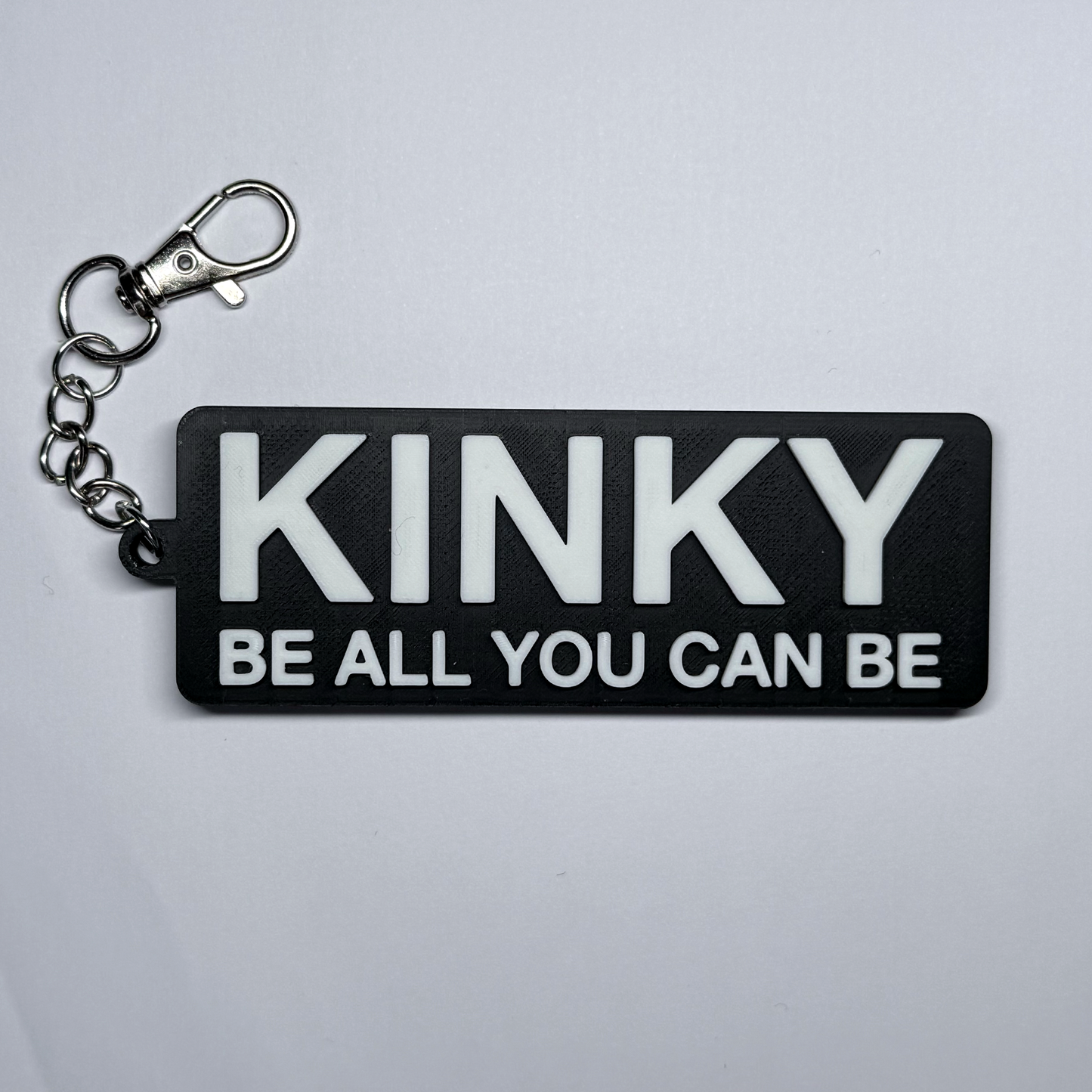 Keychain with 'KINKY BE ALL YOU CAN BE' text on a gray background